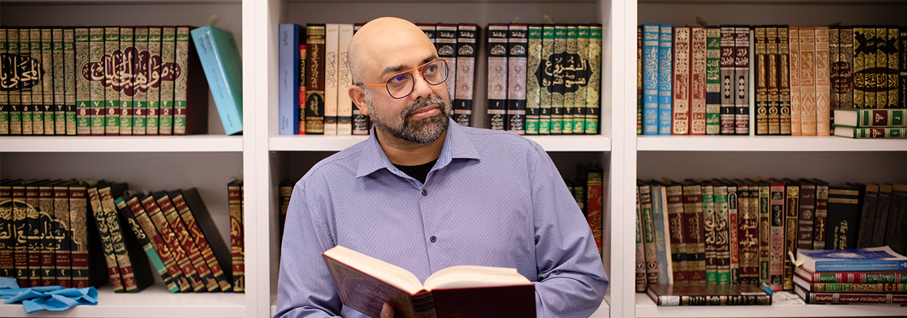 Academic man with open book standing in front of a bookcase full of Islamic books