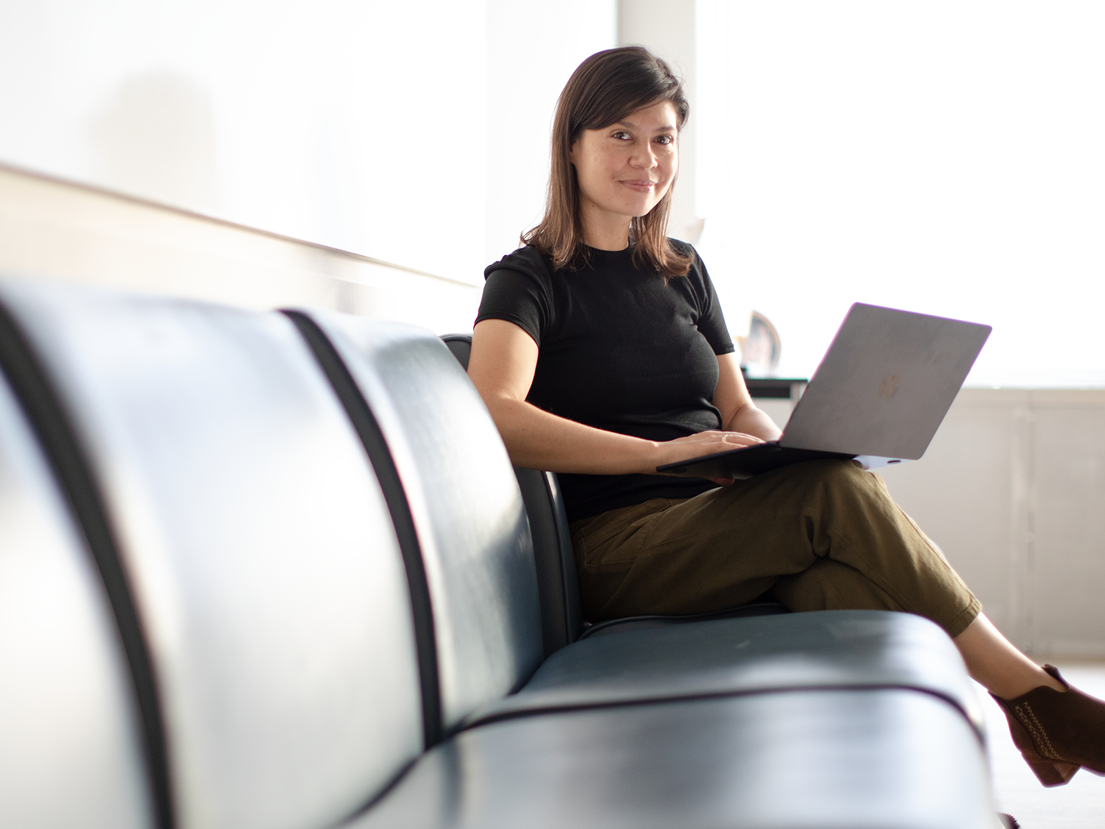 Woman in profile sitting on a bench with a laptop on her knee facing the camera.